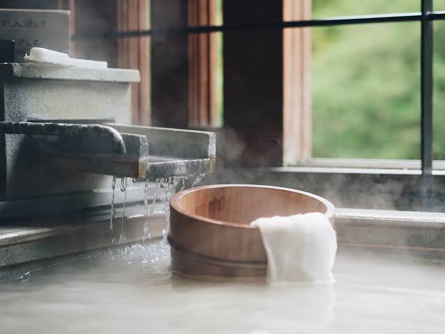 A traditional wooden bath bowl filled with steaming hot spring water, with a white towel draped over the rim and water dripping from stone channels in the background.