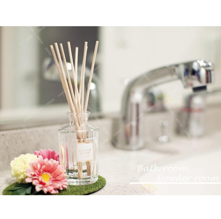 A clear glass reed diffuser bottle with natural wooden reeds, styled on a bathroom counter beside pink and yellow flowers. A blurred chrome faucet and bathroom background are visible behind it, with the text 'Bath room Powder room' overlaid on the lower right.