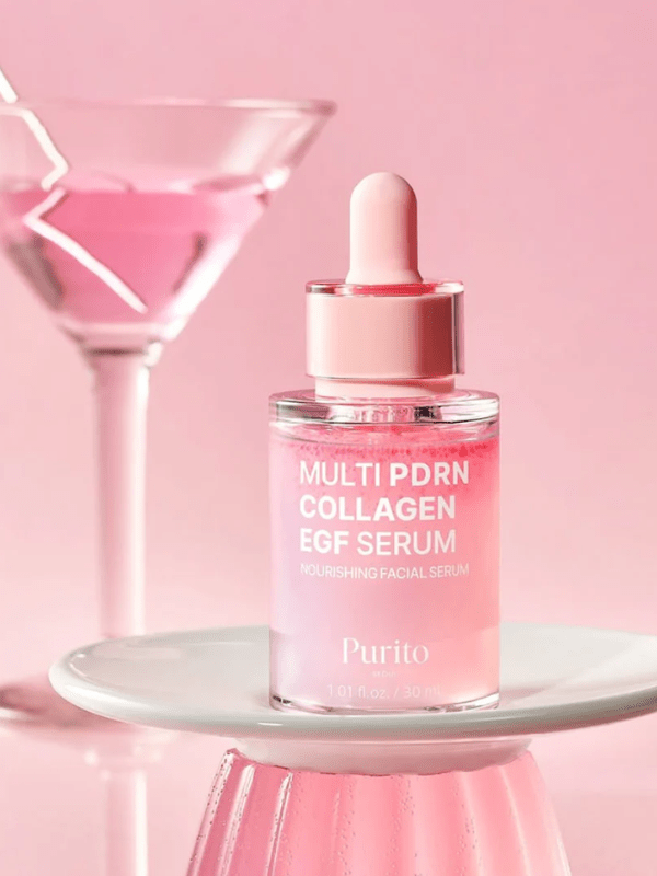 Purito Multi PDRN Collagen EGF Serum in a clear dropper bottle with pink liquid, displayed on a small white pedestal against a pink background with a blurred pink drink in a martini glass.
